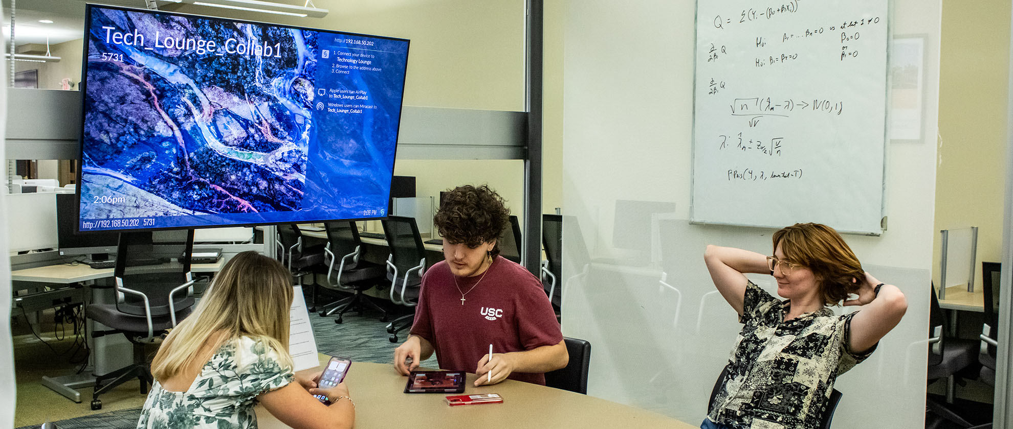 Before a Tech Lounge Collab monitor, one student is on her phone, another uses their tablet, and the third stretches their arms.