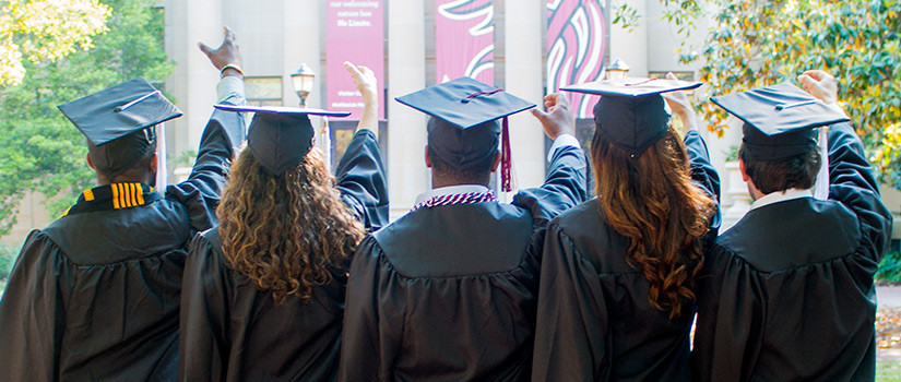 five students facing the McKissick Museum wearing black caps and gowns with their right hands raised and fingers cupped reciting UofSC alma mater