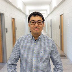 a photo of a man with a blue check button up shirt on smiling in a white hallway