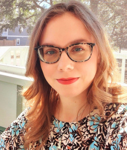 Carlie wears a floral patterened shirt and glasses and smiles at the camera