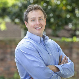 Sam wears a blue oxford shirt, is posed with his arms crossed, and stands near a brick garden wall