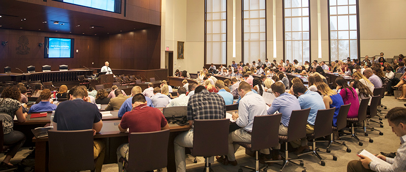 wideshot of auditorium filled with students in foreground and professor at podium in background.