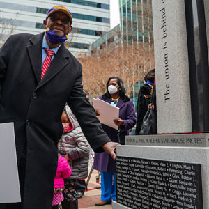 man with red tie, black coat, baseball cap standing with granite marker