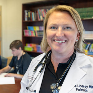 Sara Lindsey wears her white coat and smiles while a student works in the background