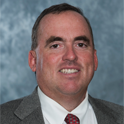 Man in a gray suit, white shirt and red tie smiling against a blue-gray studio background.
