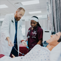 Healthcare professional guides two students as they examine a patient simulator with a stethoscope in a clinical training lab.