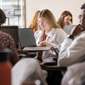 Students in white lab coats collaborate around laptops at a table.