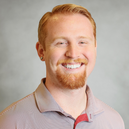 Professional headshot of a smiling person with short red hair and a neatly trimmed red beard wearing a light gray and maroon striped collared polo shirt. The person is facing the camera and is wearing a small stud earring. The photo is set against a soft gray studio background with warm, even lighting, creating a friendly, approachable, and professional appearance.