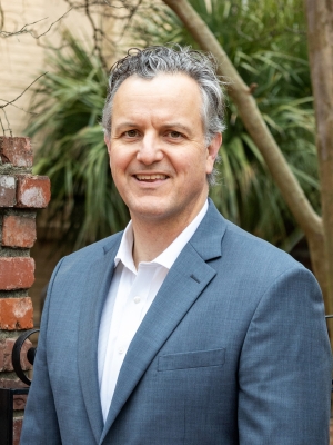 Professional headshot of Samuel Y. Todd wearing a blue blazer over a white open-collar shirt, photographed outdoors with greenery and a brick column in the background.