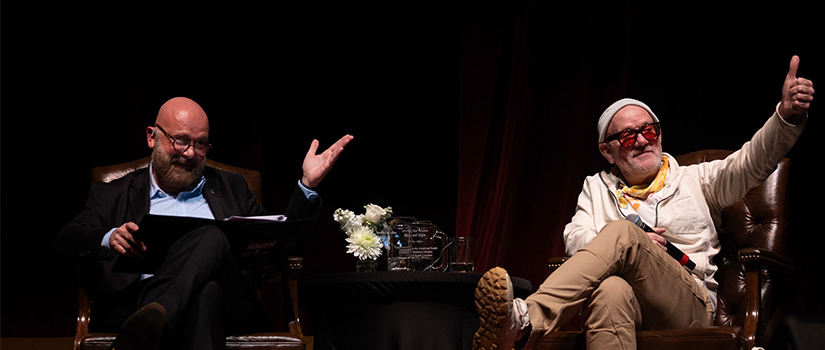 Two men seated on stage during a conversation or panel event. The man on the left, bald with a beard and wearing glasses and a dark suit, gestures with both hands while holding a folder. The man on the right, wearing a white jacket, beige pants, a white beanie, and red-tinted glasses, gives a thumbs-up with a relaxed expression. A small table with flowers, water glasses, and an award sits between them. The background is dark, focusing attention on the speakers.