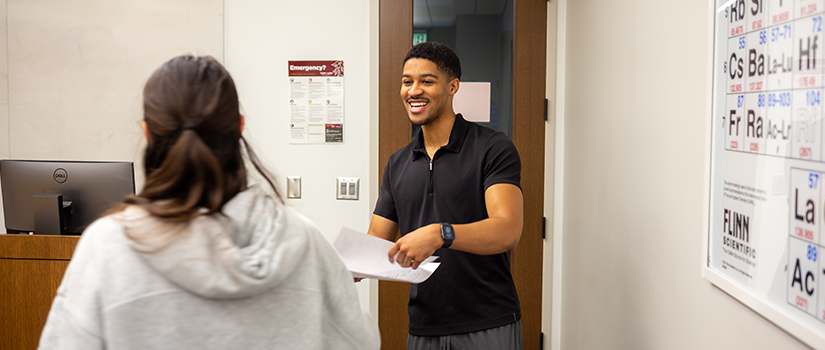 Student hands a paper to a classmate in a chemistry classroom, with a periodic table displayed on the wall nearby.