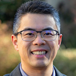 Portrait of a man wearing glasses, smiling, outdoors with a blurred background.