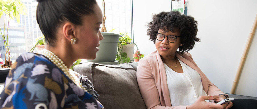 Two women of color sit on a gray couch in a business office setting. They are turned slightly to face each other in conversation and their expressions are friendly.