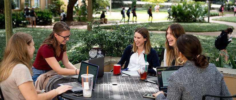 A group of graduate students sit at a table outside of the psychology building. They are talking and laughing, and some are working on laptop computers.