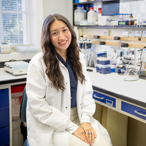 Woman in white lab coat sits at a table with lab equipment.