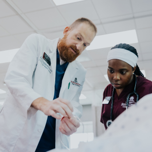 teacher in lab coat helping nursing student in scrubs place an iv on a mannequin